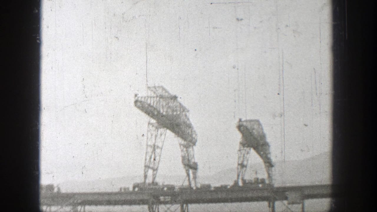 Two cranes stand over a bridge structure in hazy black-and-white footage.