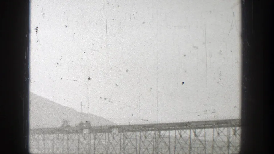 1938 Washington 8mm view of a train or rail cars moving across a long bridge span with hillside terrain behind it.