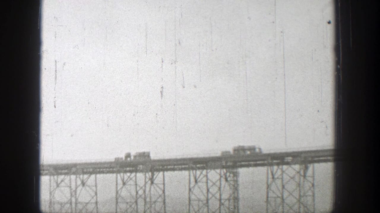 1938 Washington home-movie frame showing small rail vehicles moving across a distant steel bridge under a pale sky.
