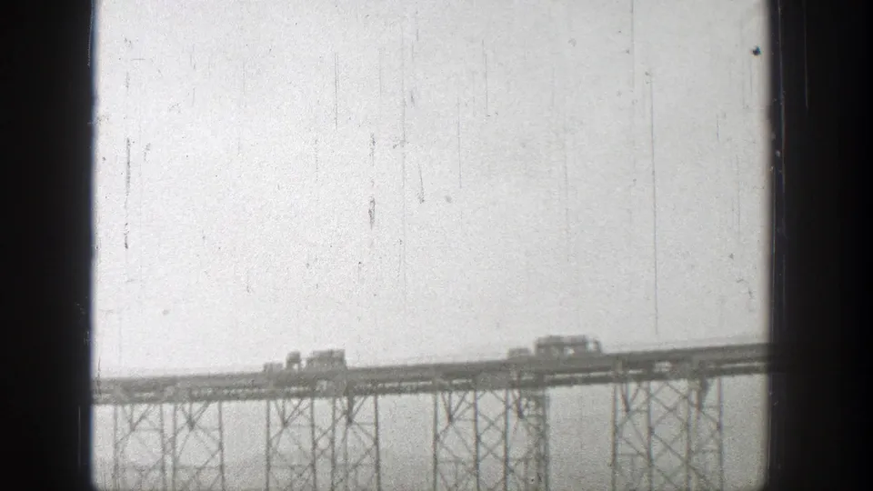 1938 Washington home-movie frame showing small rail vehicles moving across a distant steel bridge under a pale sky.