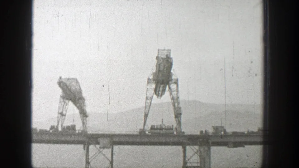 1938 Washington home-movie frame showing large bridge cranes and small vehicles on an elevated span against hazy hills.