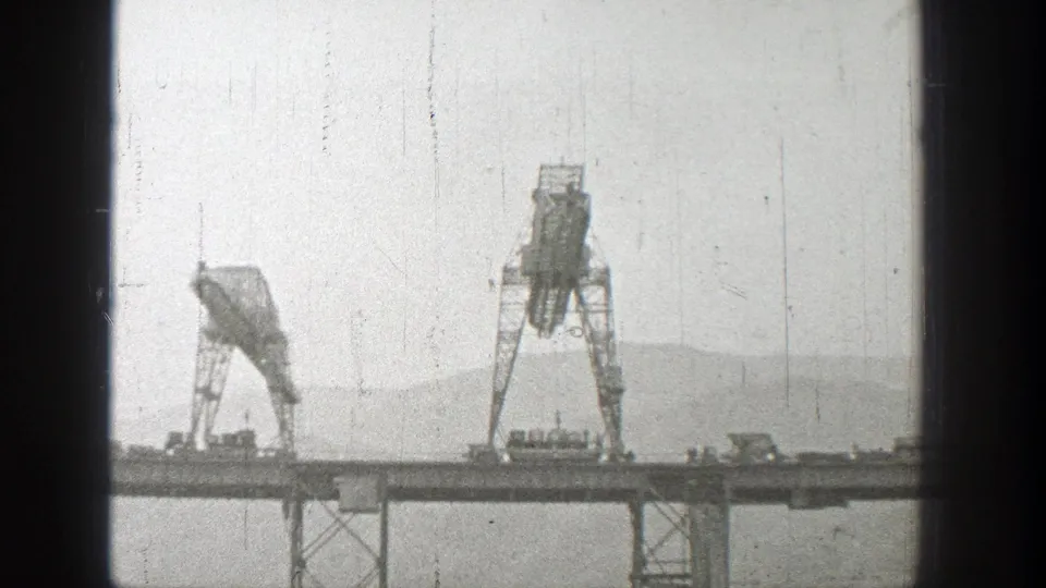 1938 Washington 8mm frame showing two gantry cranes on a bridge under construction, with water and hazy hills behind.