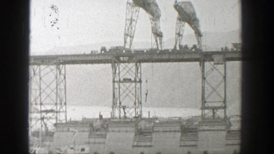 Late-frame view of the 1938 Washington bridge construction scene, with cranes above steel supports and water in the background.