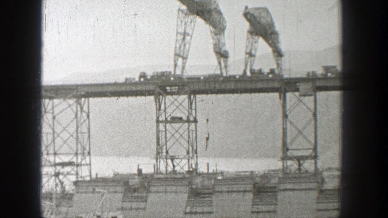 1938 Washington 8mm frame showing two cranes on unfinished bridgework above water, with scaffold towers and hazy hills beyond.