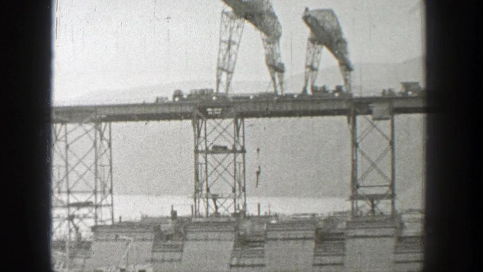 1938 Washington 8mm frame showing two cranes on unfinished bridgework above water, with scaffold towers and hazy hills beyond.