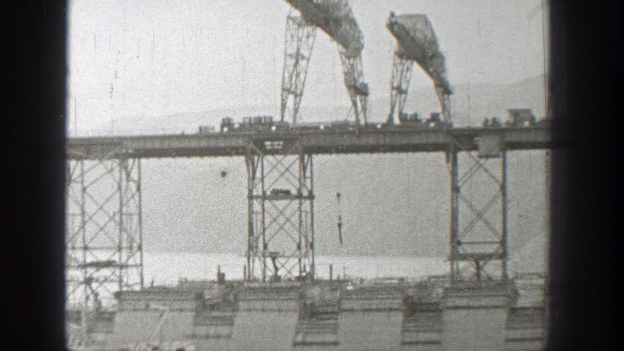 Bridge construction scene with two cranes, a lowered hook, water, and hazy hills.