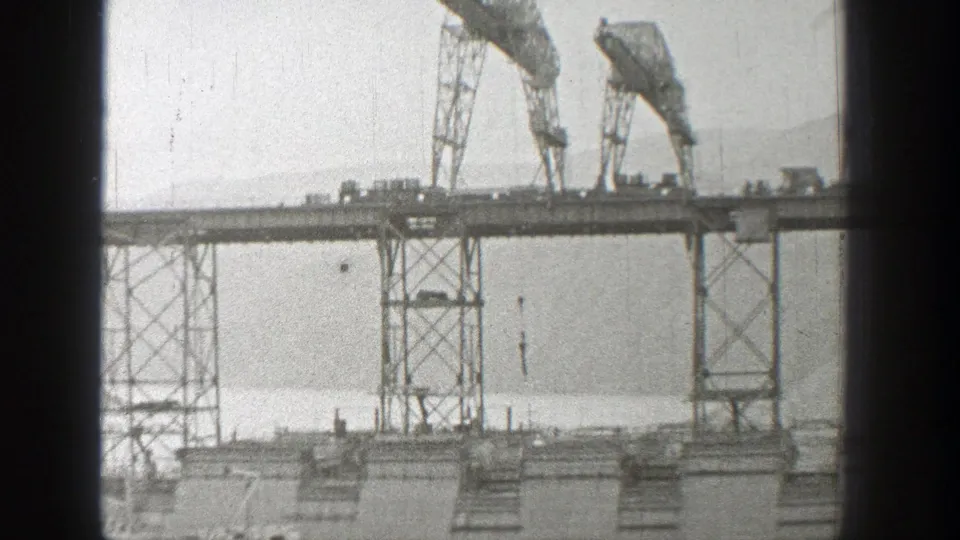 1938 Washington bridge construction view with twin cranes on the span and a hook hanging over the water below.