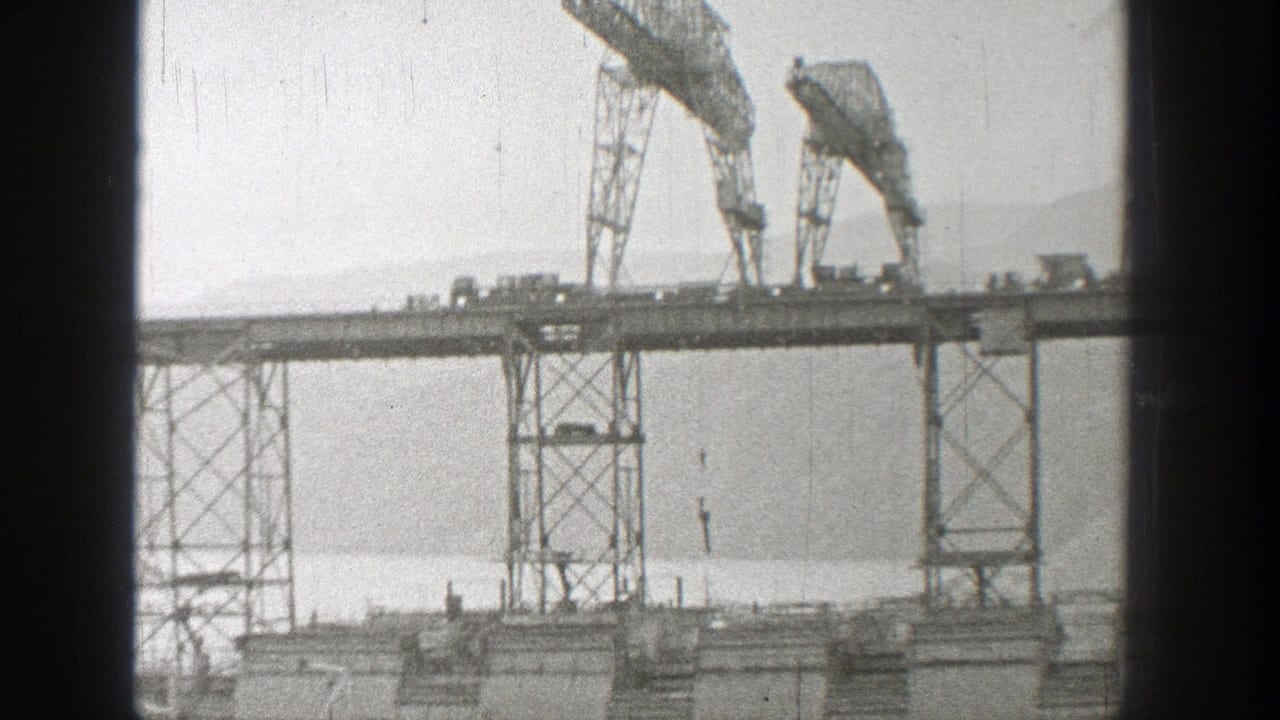 1938 Washington 8mm frame showing a high trestle bridge under construction, with two gantry cranes, small vehicles, water, and distant mountains.