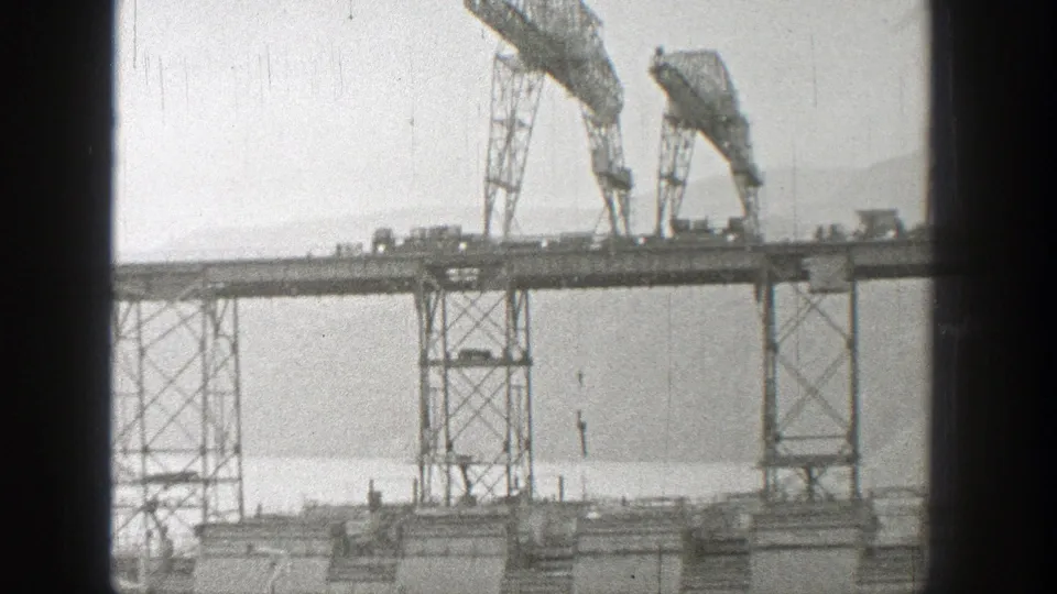 1938 Washington 8mm frame showing a high trestle bridge under construction, with two gantry cranes, small vehicles, water, and distant mountains.