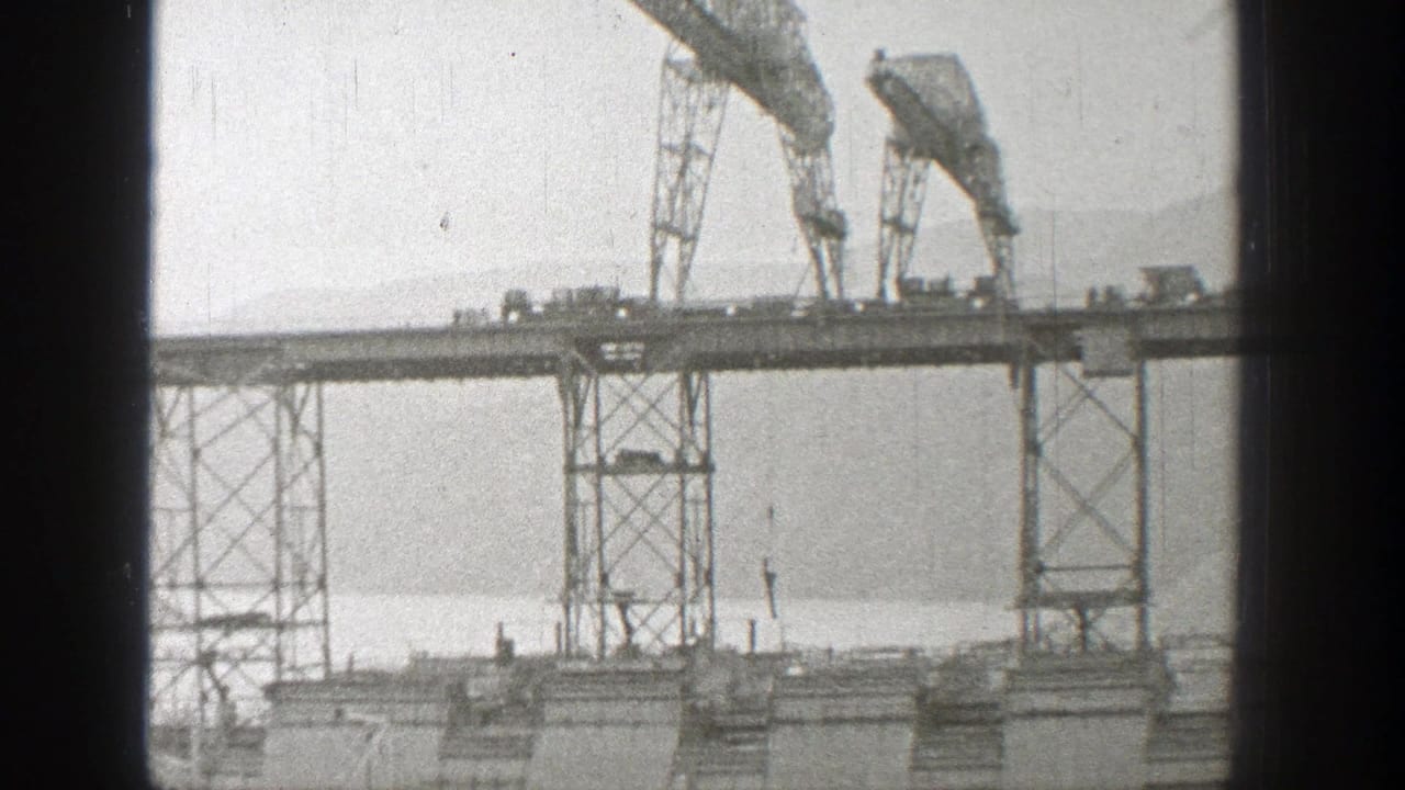 Trestle bridge span with cranes, vehicles, water, and a mountain backdrop.