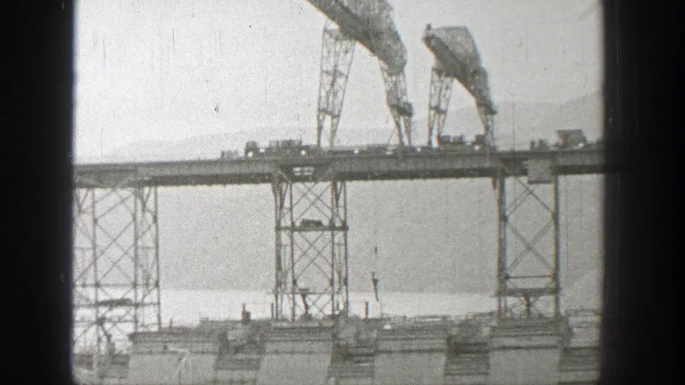 1938 Washington home-movie frame of a steel trestle bridge span, gantry cranes, small vehicles, open water, and hazy mountains.