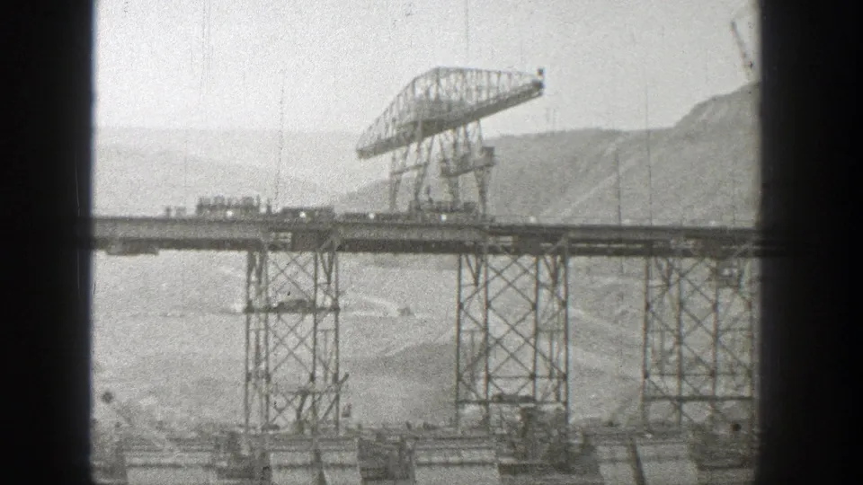 1938 Washington frame showing a large gantry crane over the trestle bridge, with lattice supports, a small vehicle, water, and hills beyond.