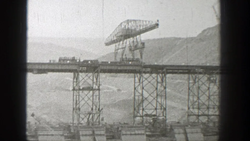 1938 Washington 8mm frame with a gantry crane on an elevated trestle bridge, open water below, and mountainous terrain in the distance.