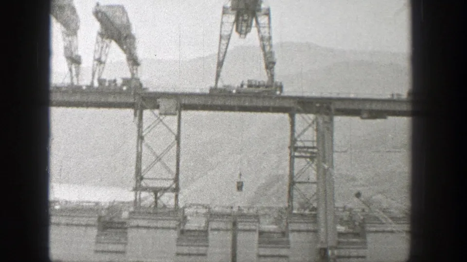 Cranes work along a steel bridge span in Washington, with suspended material, water, and hills visible behind them.