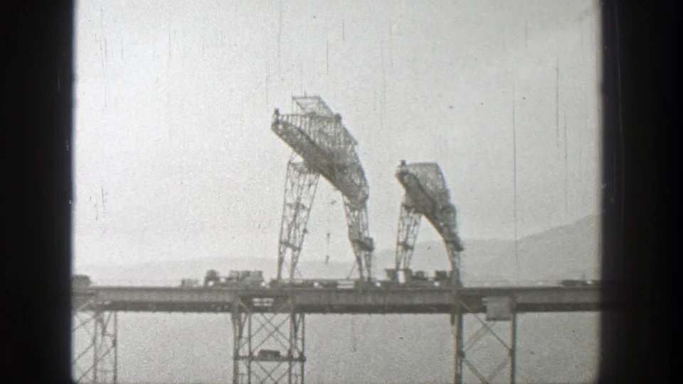 Two large gantry cranes stand on a Washington bridge construction span, framed by pale sky, hills, and 8mm film edges.