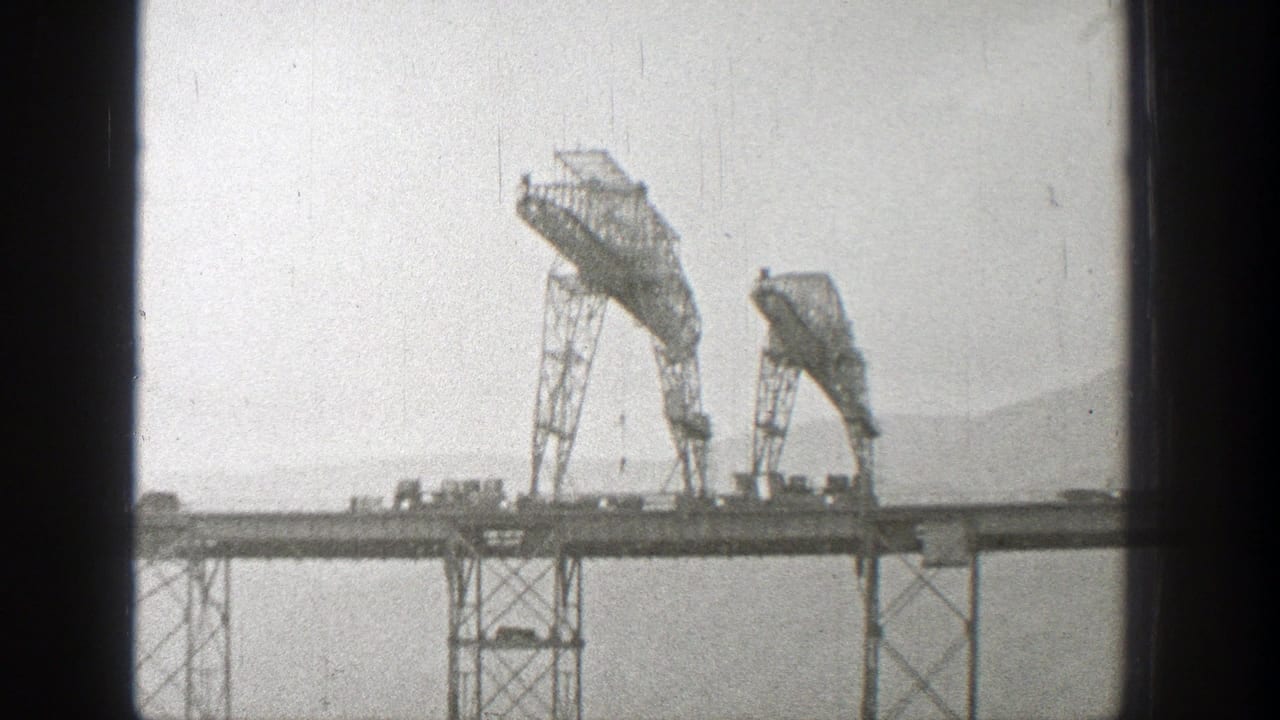 Steel truss bridge with two tall metal sections, small vehicles, and pale hillside background.