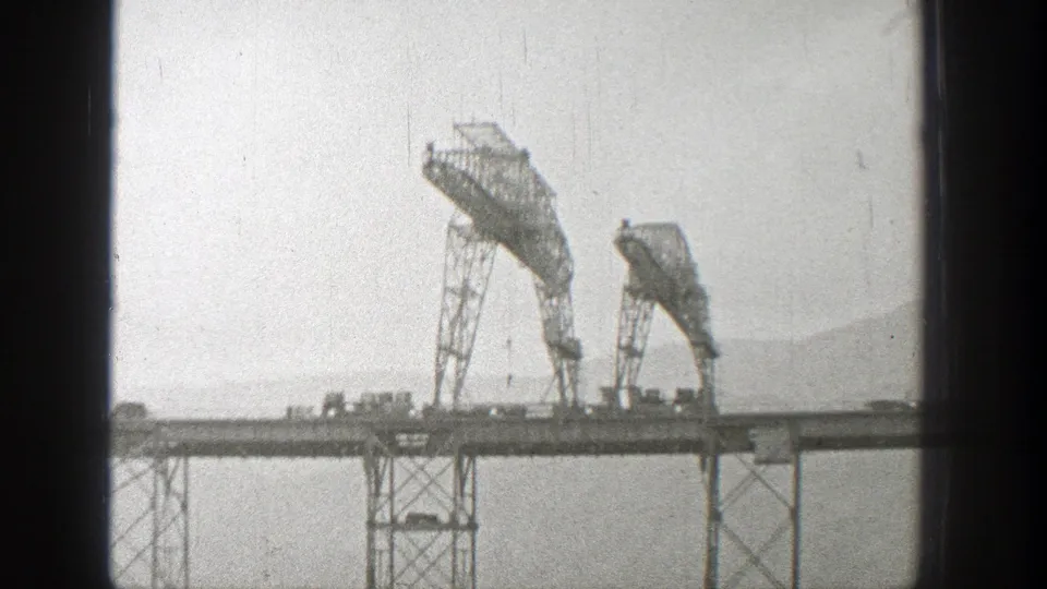 Washington home-movie frame from 1938 showing the steel bridge structure, roadway traffic, pale sky, and distant hillside through grainy 8mm film.