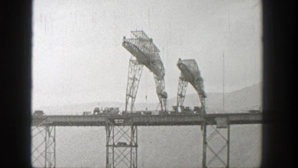 Black-and-white 1938 Washington frame of a steel bridge with lattice supports, two raised metal sections, light traffic, and hazy hills beyond.