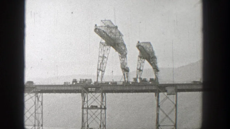 Final 1938 Washington 8mm frame showing the steel truss bridge, two prominent metal sections, small vehicles, haze, and distant hillside.
