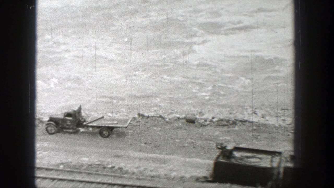 Flatbed truck and abandoned car body beside rail tracks near a rough riverbank.