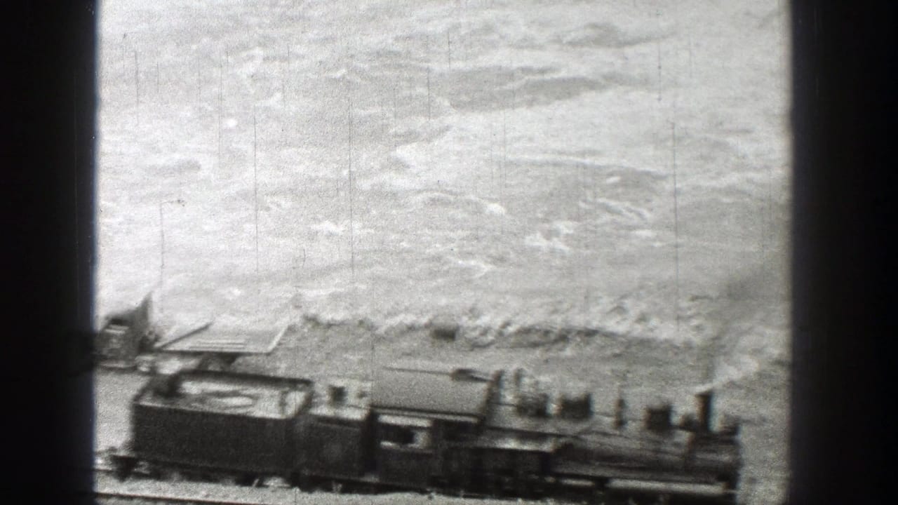 Steam locomotive passing along tracks beside the riverbank and parked vehicle.