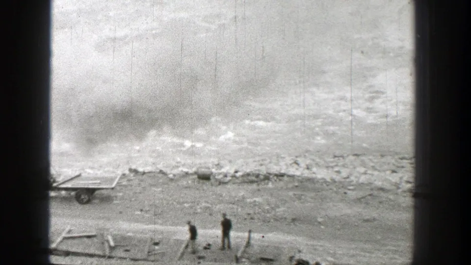 Two distant people stand by the riverside tracks in 1938 Washington, with smoke or haze over the rough riverbank.
