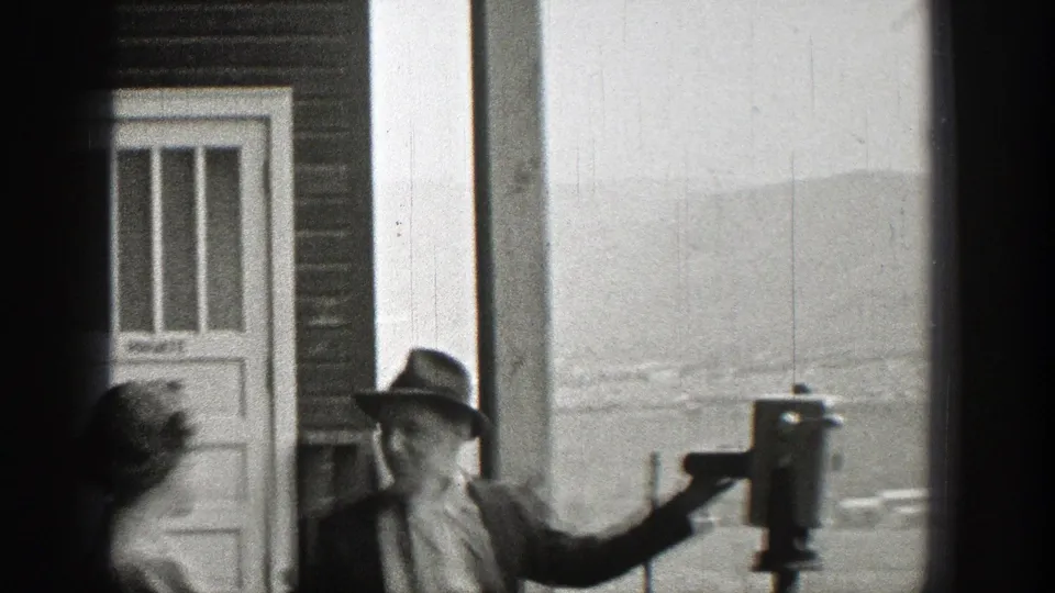 In 1938 Washington home-movie footage, a suited man in a fedora stands by a coin-operated viewing scope as a woman passes nearby.