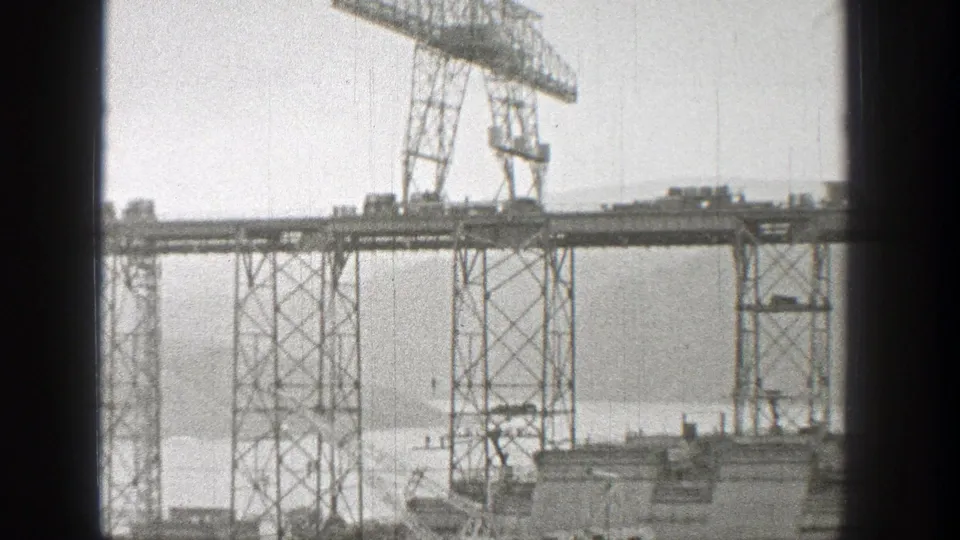 Steel bridge construction in Washington, 1938, with a gantry crane and lattice supports silhouetted against water and distant land.