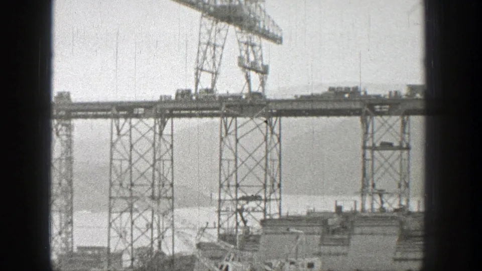 Distant 8mm view of a bridge construction deck, steel towers, and gantry crane over water in Washington in 1938.