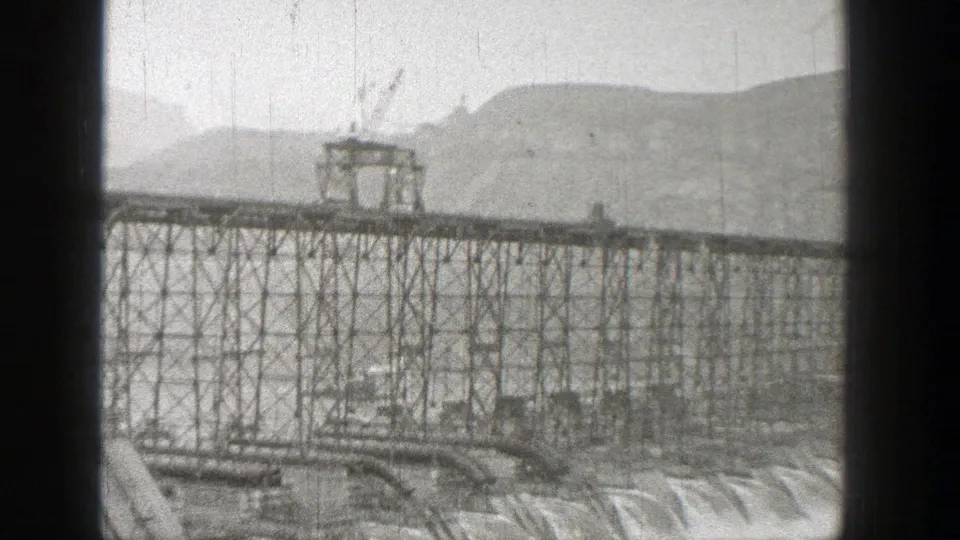 1938 Washington 8mm frame of a truck moving across an elevated steel bridge over water, with hills in the background.