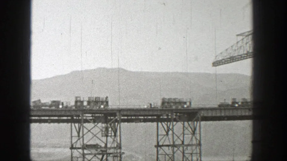 1938 Washington 8mm frame of two trucks crossing a steel bridge span high above water, backed by distant hills.