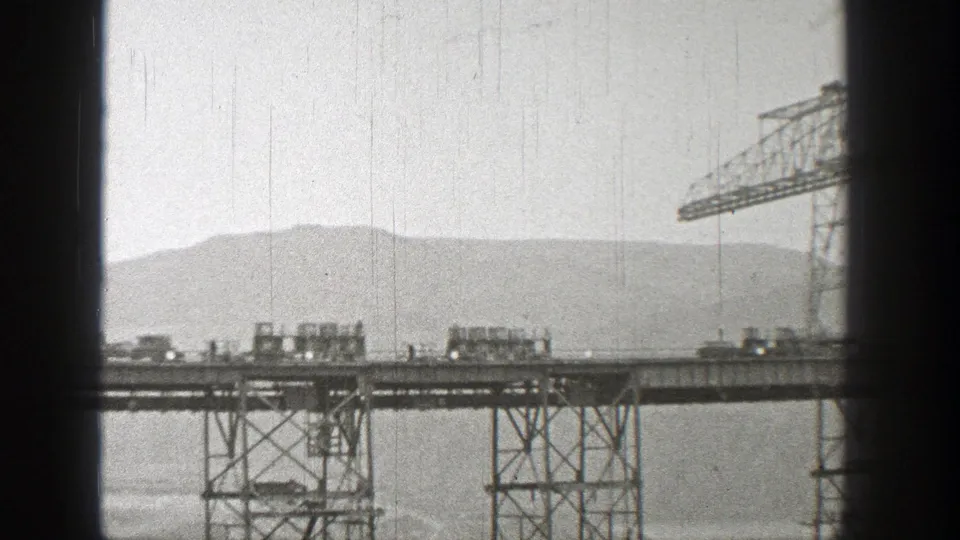 1938 Washington 8mm frame showing rail cars on a tall steel bridge with construction supports and distant mountains.