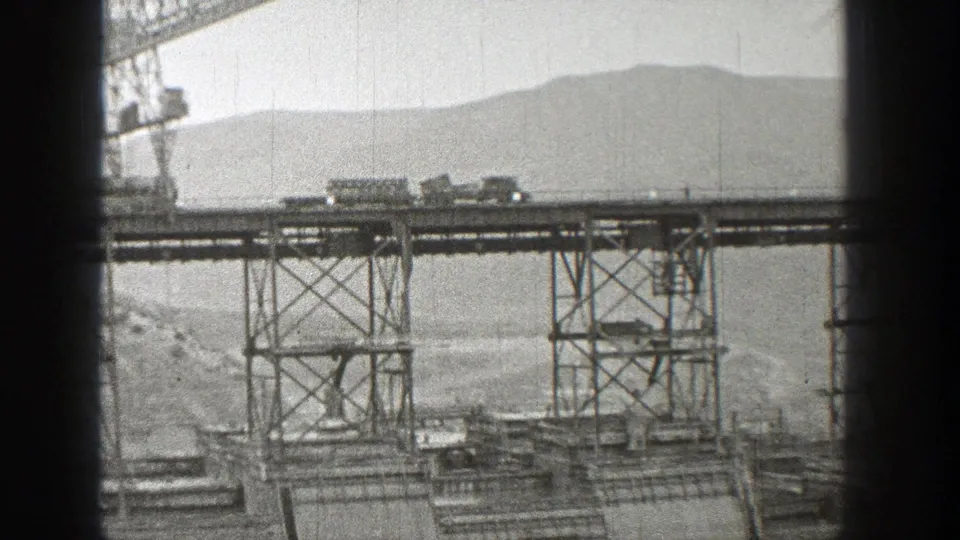 Black-and-white 1938 Washington frame of train cars on an elevated steel bridge beside crane and support structures.