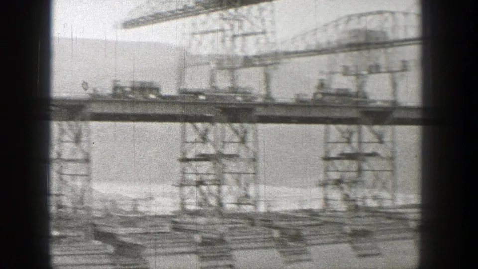 Soft black-and-white 1938 Washington frame of a train crossing a steel bridge amid cranes, scaffolding, and distant hills.