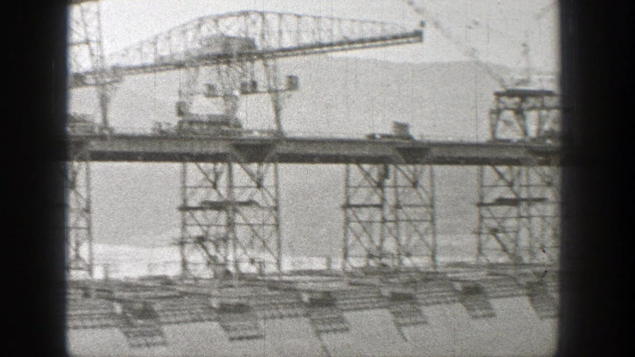 1938 Washington 8mm frame showing steel towers and crane structures crossing a dam construction site against a mountain backdrop.