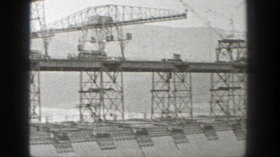 1938 Washington 8mm frame showing steel towers and crane structures crossing a dam construction site against a mountain backdrop.