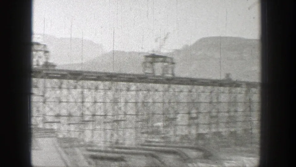 Wide 1938 Washington view of dam construction, with lattice towers, elevated crane tracks, and mountain slopes in the distance.