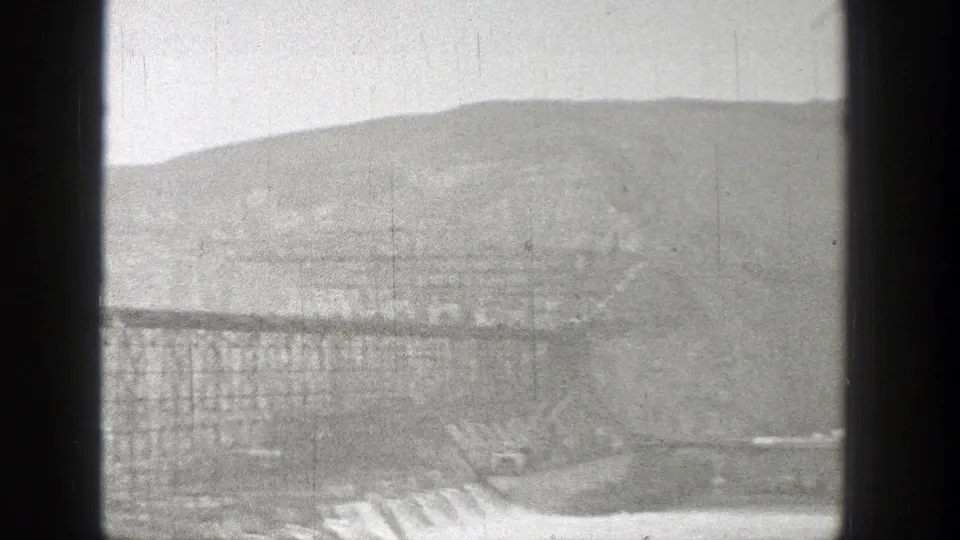 Wide 1938 Washington frame of a dam worksite, showing spillway flow, steel scaffolding, and hazy mountain terrain.