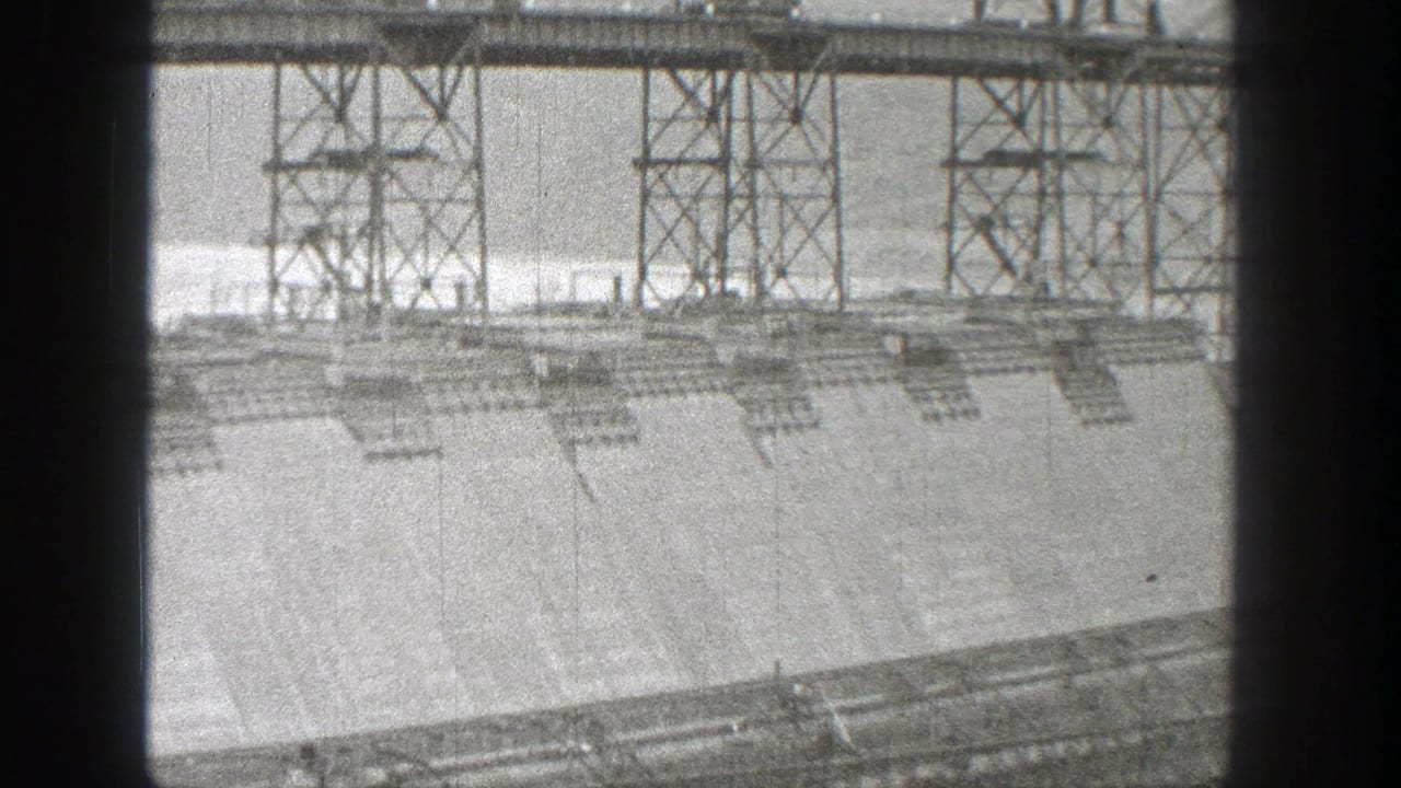 Concrete dam face with steel support towers and water beyond.
