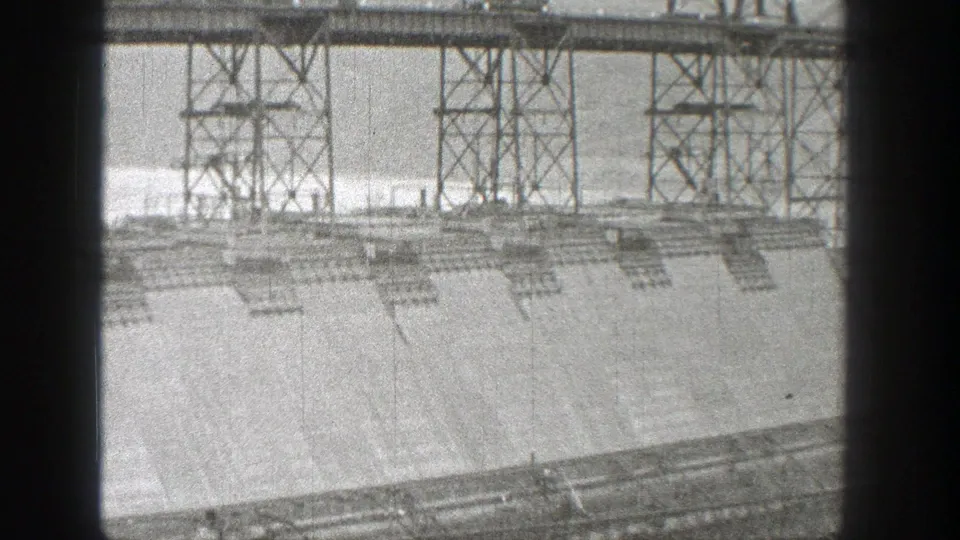 1938 Washington dam construction view with steel lattice towers above a broad concrete face and reservoir water in the background.
