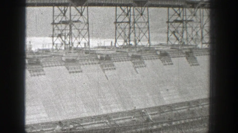 Distant view of a Washington hydroelectric dam under construction, showing steel tower supports and the sloped concrete face.