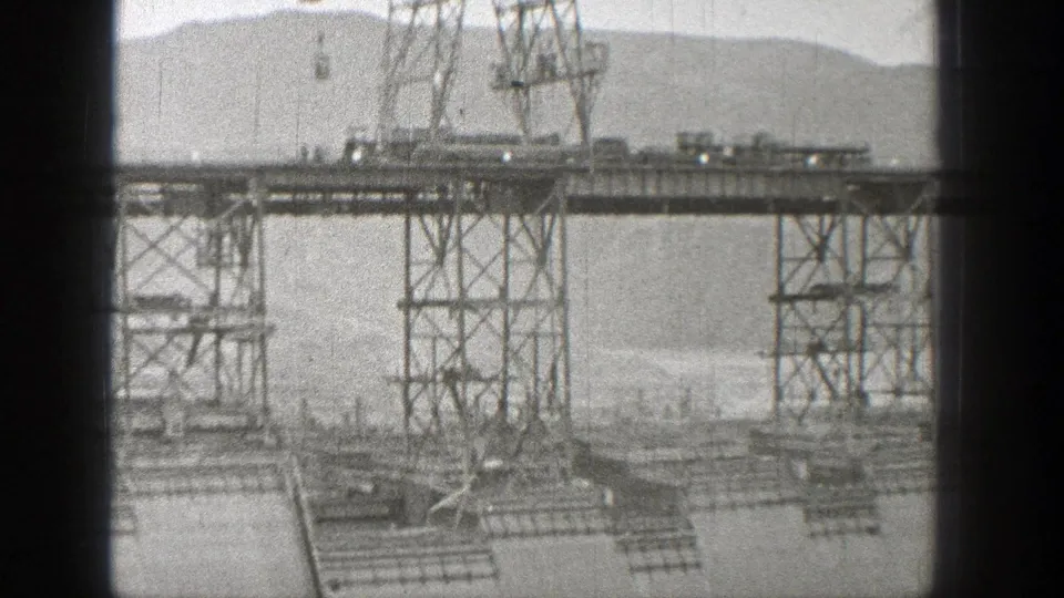 Washington dam construction scene with steel lattice towers, platforms, distant workers, concrete surfaces, water, and hills beyond.