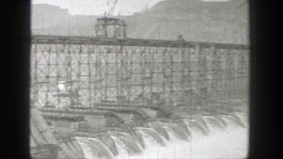 1938 Washington frame of a dam spillway with a steel truss roadway and small vehicles visible above the water release.