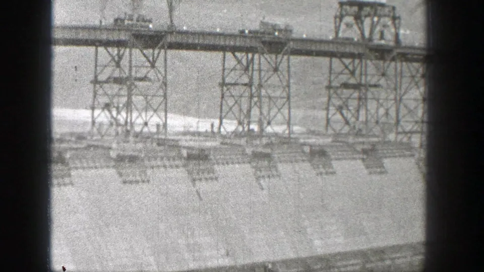 Black-and-white 8mm view of a steel-supported roadway over a Washington dam, with spillway water and concrete structures below.