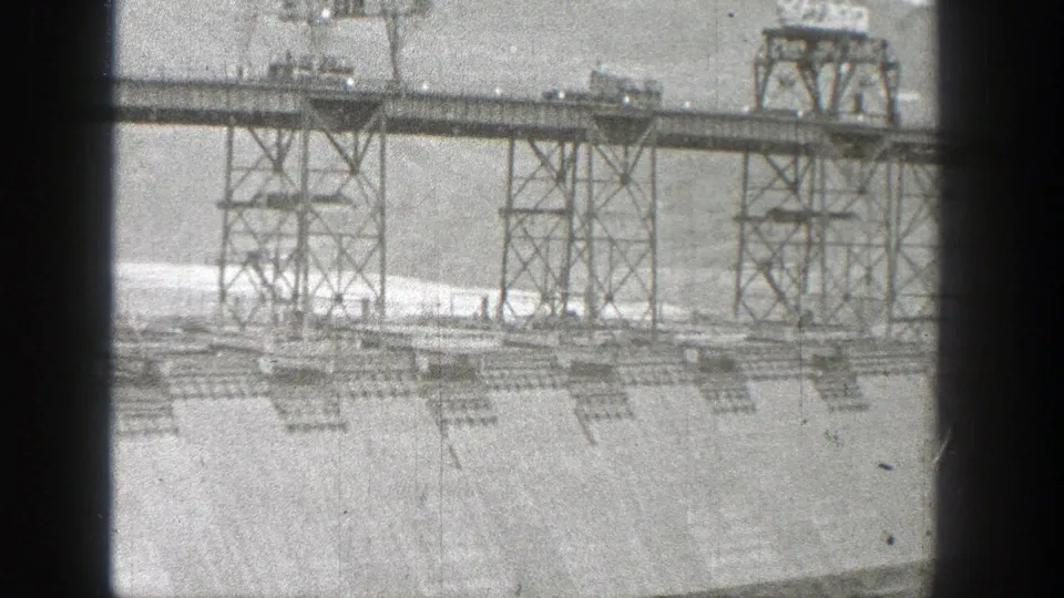 1938 Washington dam footage showing a steel truss roadway with vehicles above a concrete spillway and pale water below.