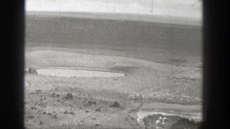 Black-and-white 8mm frame of a dry Washington riverbed with pale exposed ground, shallow water, and scattered stones.
