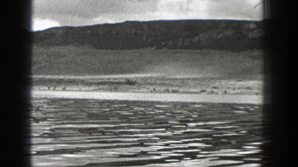 1938 Washington 8mm frame looking across rippling water toward a low, dark ridge and pale shoreline.