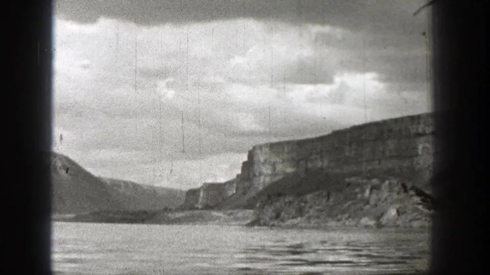 Black-and-white frame of a Washington lakeside landscape with calm water, rocky cliffs, distant slope, and visible film wear.