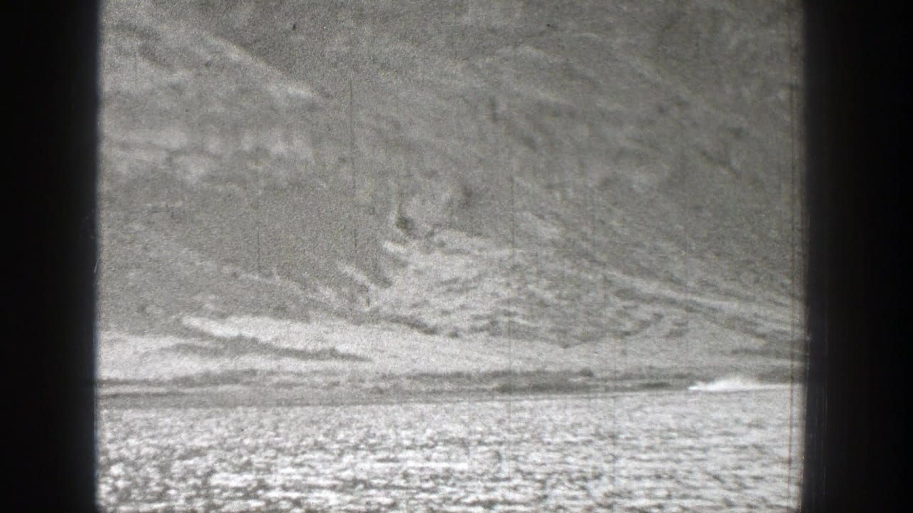 1938 Washington 8mm frame showing a distant small craft or aircraft moving across a flat strip beneath textured hills, with smoke visible behind it.