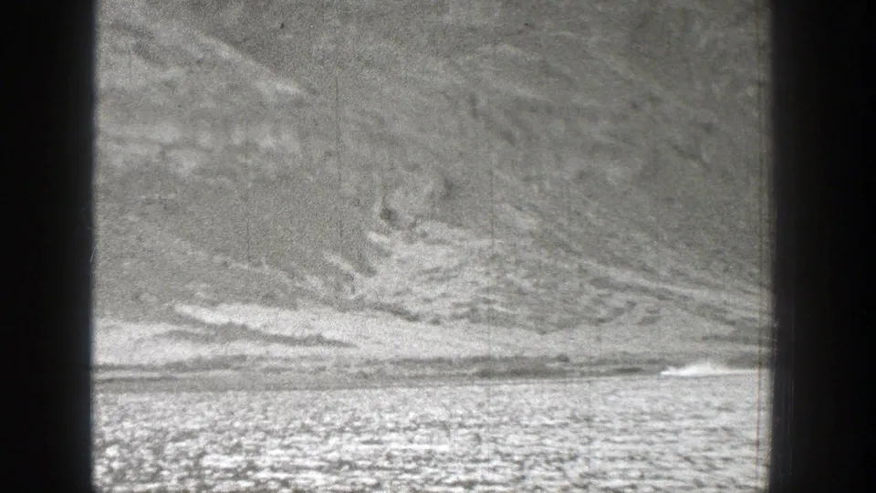 1938 Washington 8mm frame showing a distant small craft or aircraft moving across a flat strip beneath textured hills, with smoke visible behind it.